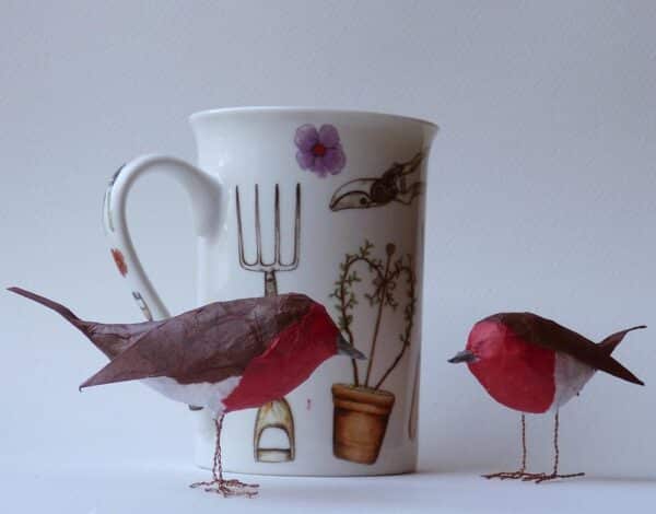 2 tiny papier mâché robins, next to a china mug